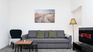 Un salon avec un canapé gris, une petite table en bois, un fauteuil vert, un lampadaire et un téléviseur affichant le logo de Netflix.