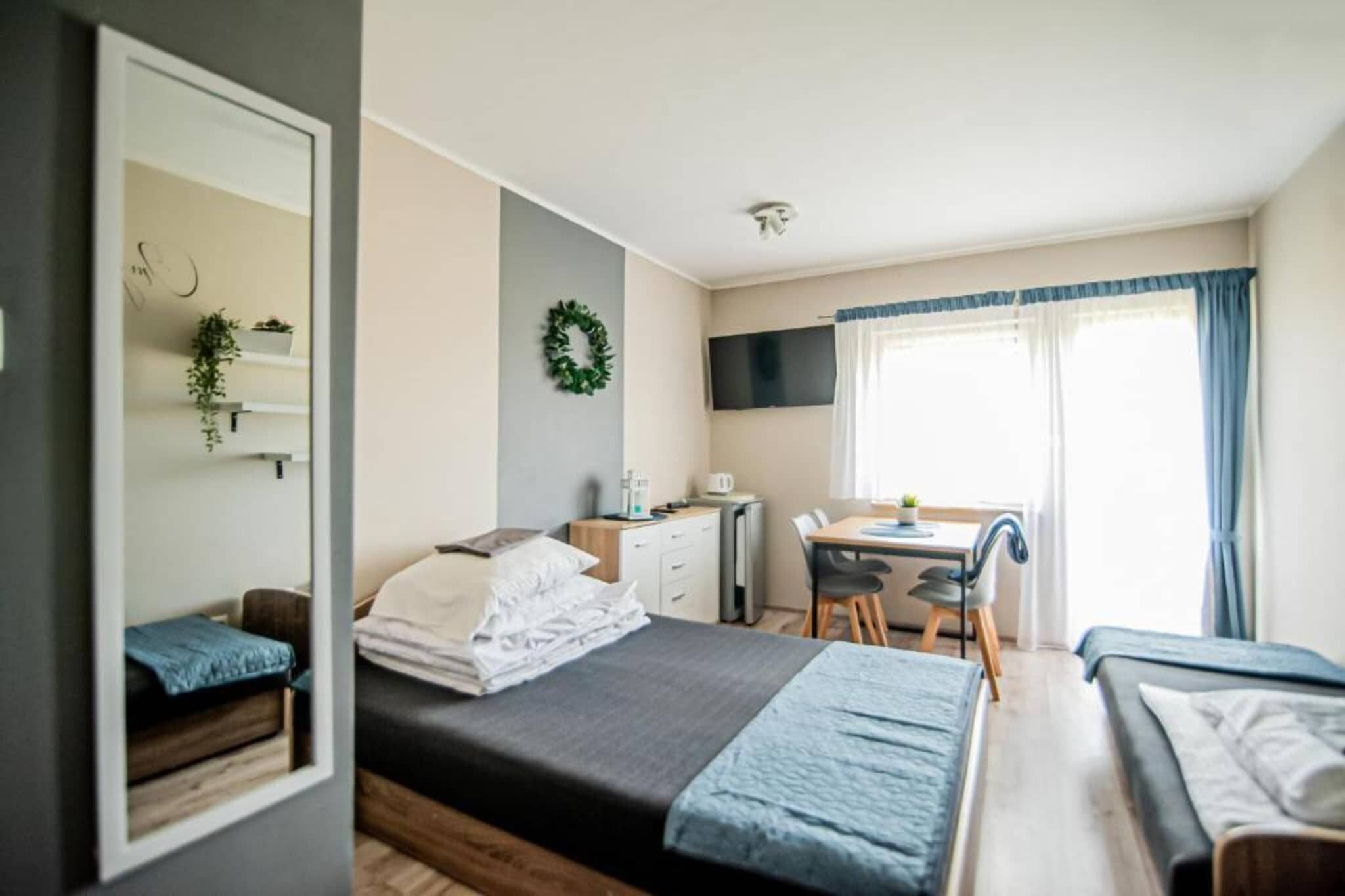 Ein kleines Zimmer mit einem Bett, einem Schreibtisch und einem Stuhl. Ein Fernseher ist an der Wand befestigt und es gibt ein Fenster mit Vorhängen.