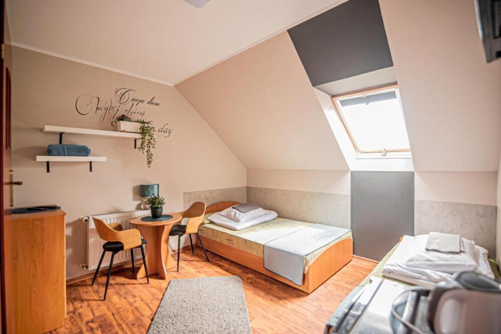 Habitación pequeña y luminosa con techo inclinado, cama, escritorio con sillas y una pequeña estantería con plantas.