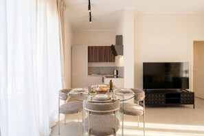 A modern living room with a glass dining table, grey upholstered chairs, a flat-screen TV, and a kitchen area with a built-in oven and microwave. - ALL Gargasole | Luxury Sky-High Retreat with 2 Private Terraces (Bari)