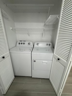 A laundry room with two stacked washers and dryers, a metal shelf above, and a tiled floor. - Beautiful Orlando Home 3BR/2BA 20 Min to Disney/Universal+ Community Pool (Orlando)
