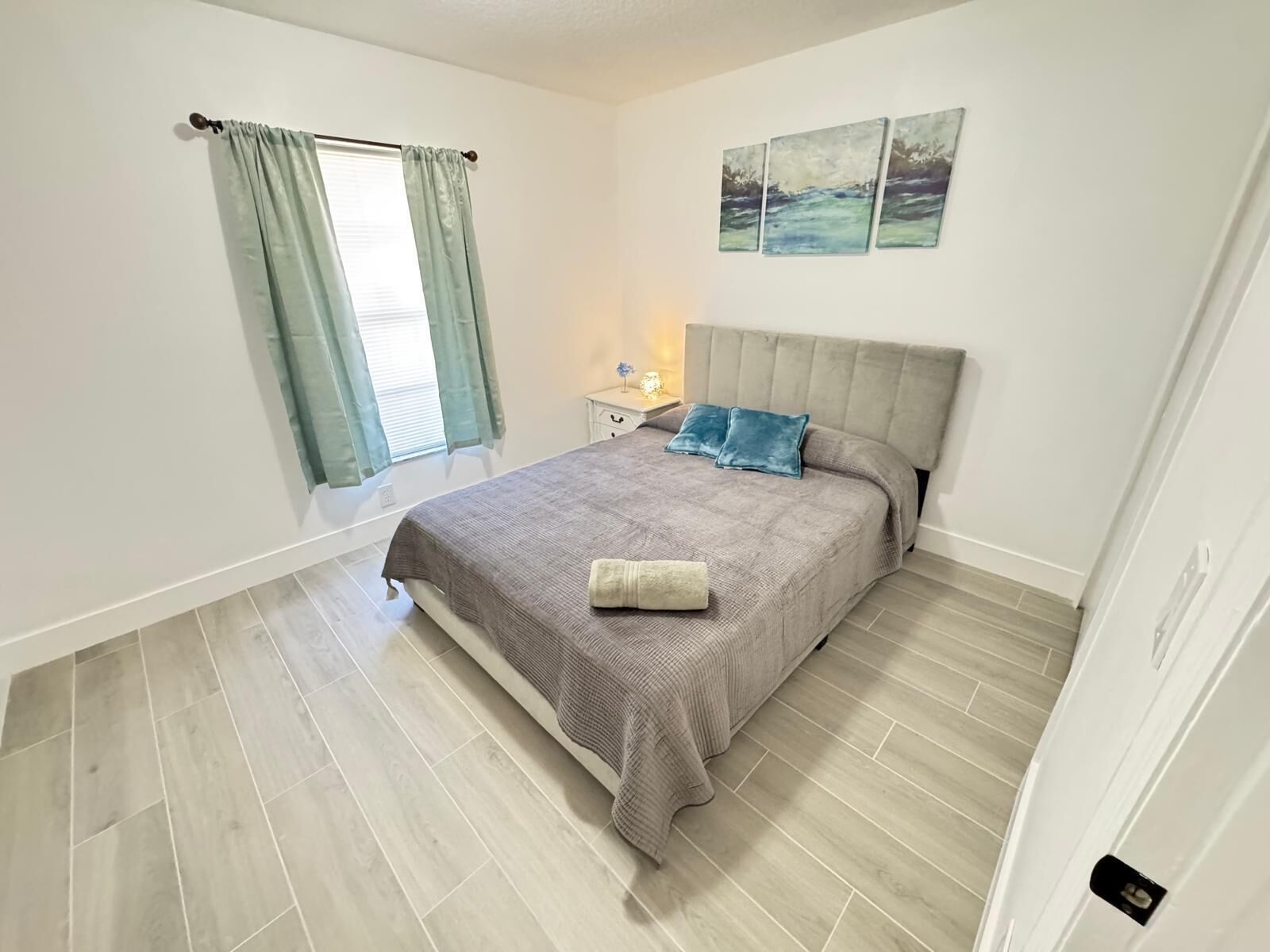 Une chambre à coucher avec un lit, une table de chevet, une fenêtre avec des rideaux et un mur orné de trois tableaux.