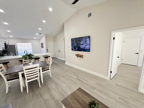 Une cuisine moderne avec des meubles blancs, un coin repas avec une table et des chaises en bois, un téléviseur fixé au mur et un couloir avec une porte.