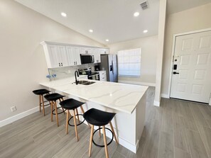 Une cuisine moderne avec des meubles blancs, un grand îlot central doté de tabourets, des appareils en acier inoxydable et une fenêtre avec des stores.