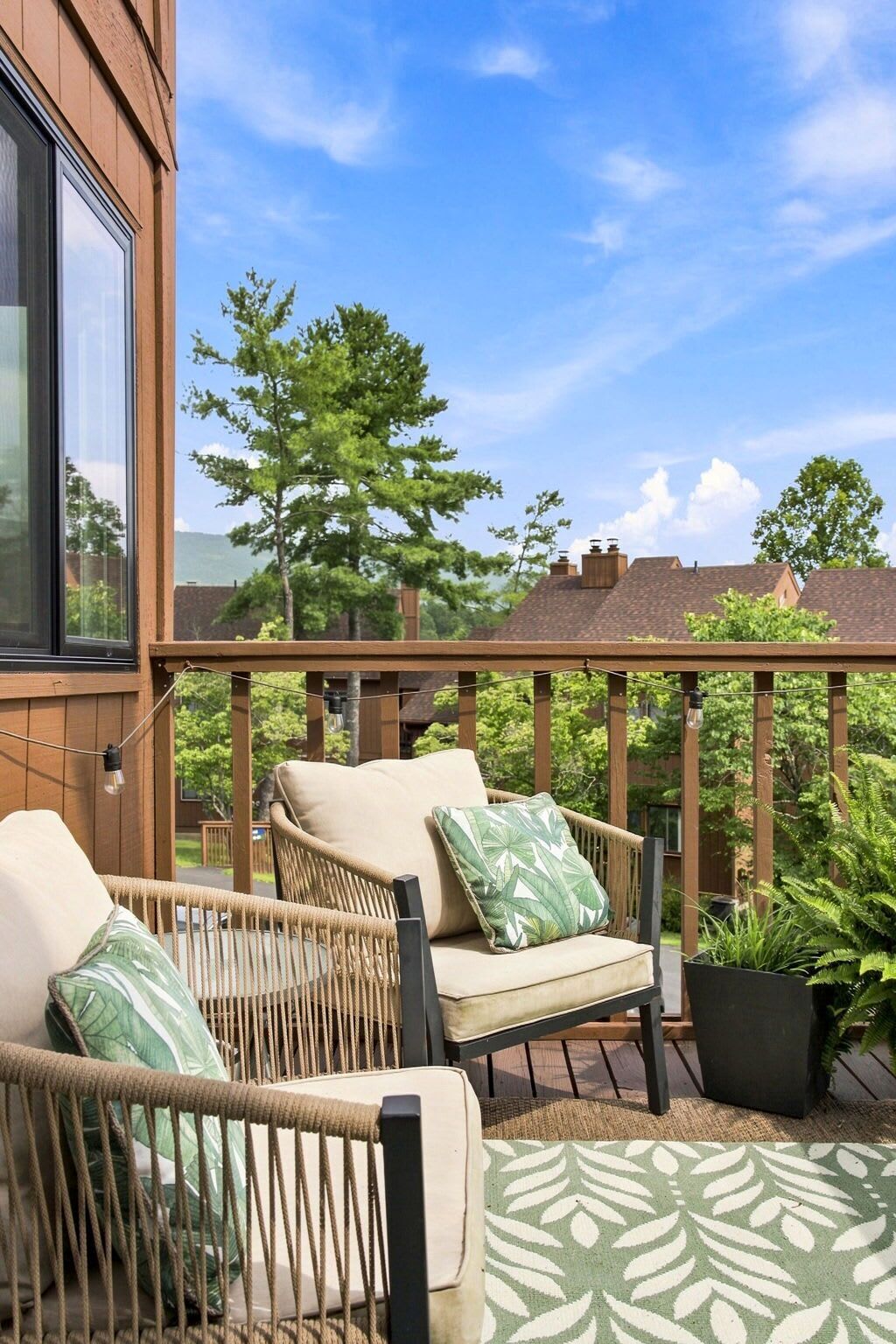 Eine Holzterrasse mit Korbstühlen und Kissen, ein Blumentopf und im Hintergrund Häuser und Bäume.