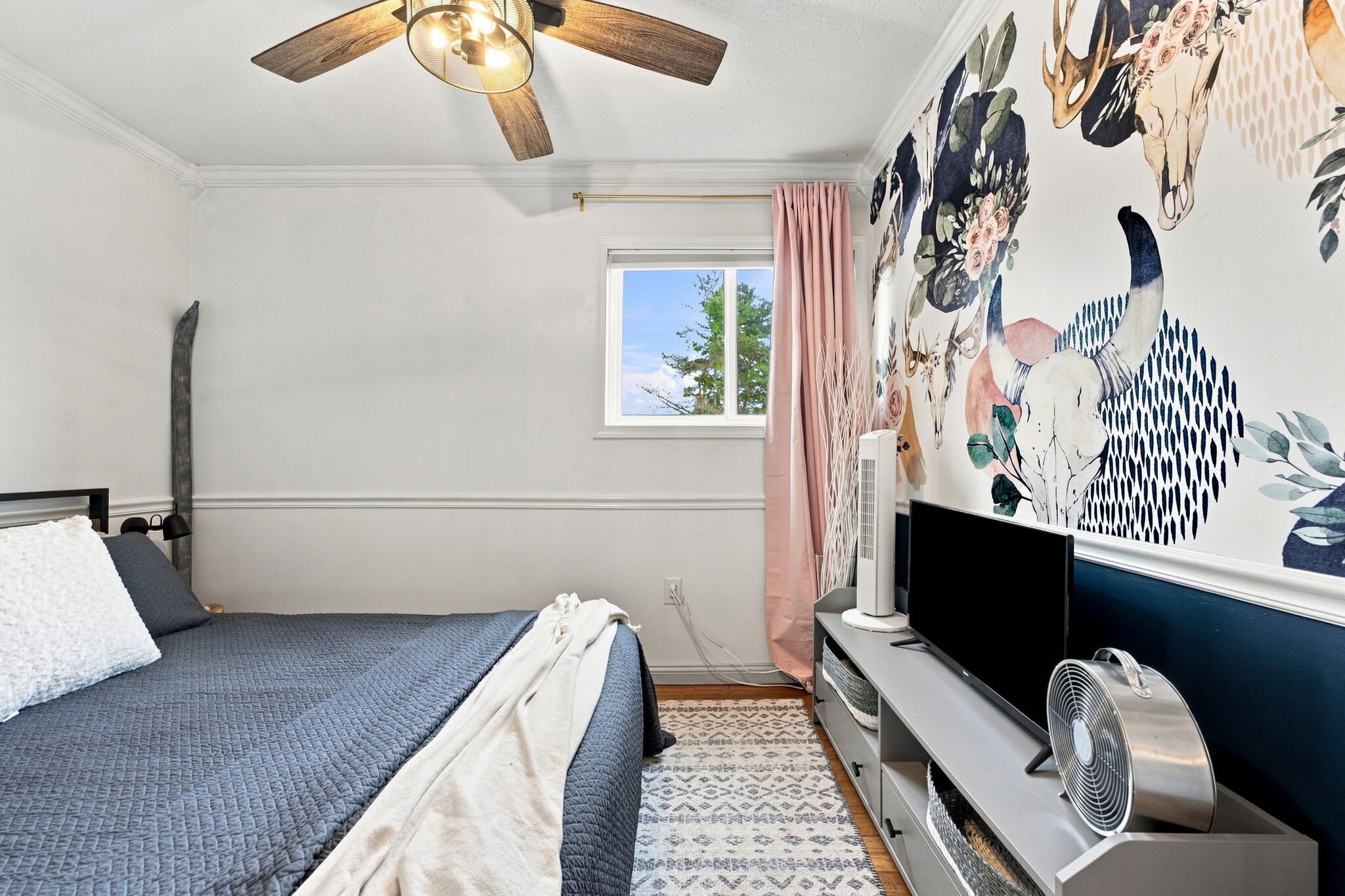 A bedroom with a bed, a ceiling fan, a window with curtains, a wall-mounted TV, and a radiator.