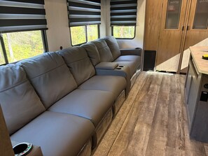 A modern RV interior with a sectional sofa, a kitchenette, and large windows. - Mountain Hideaway - 2 br 1.5 ba RV (Watauga, TN)