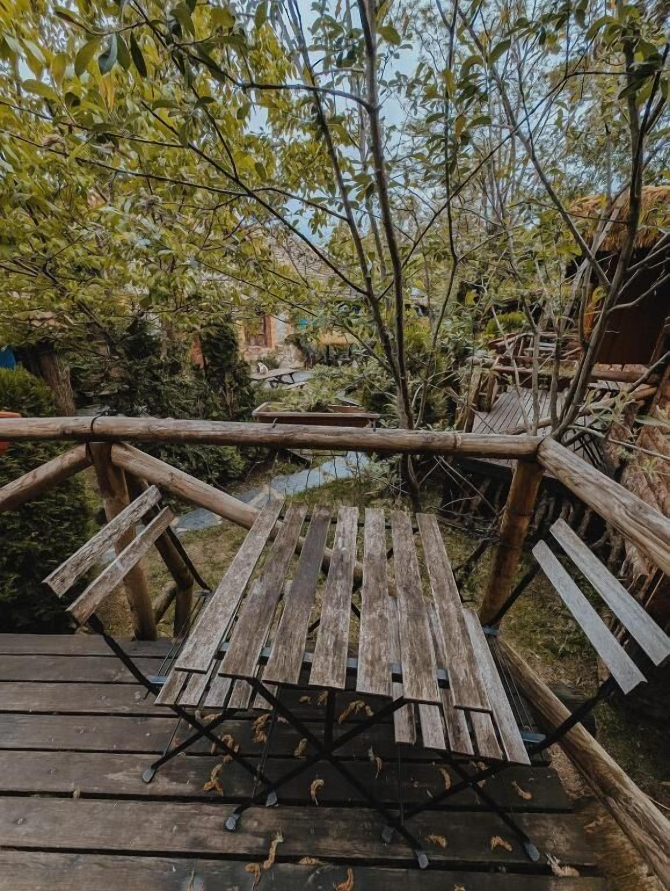 Una terraza de madera con una mesa y bancos, rodeada de árboles y una construcción con techo de paja.