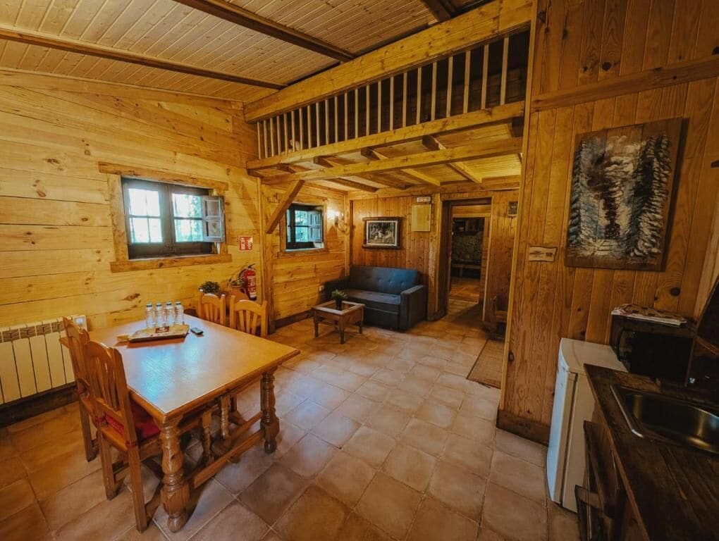 Interior de una cabaña de madera con una mesa de comedor, sillas, un sofá y una zona de cocina.