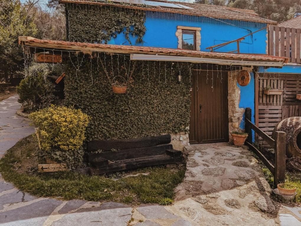 Un edificio rústico con fachada azul, un banco de madera y un sendero de piedra que lleva a la entrada.