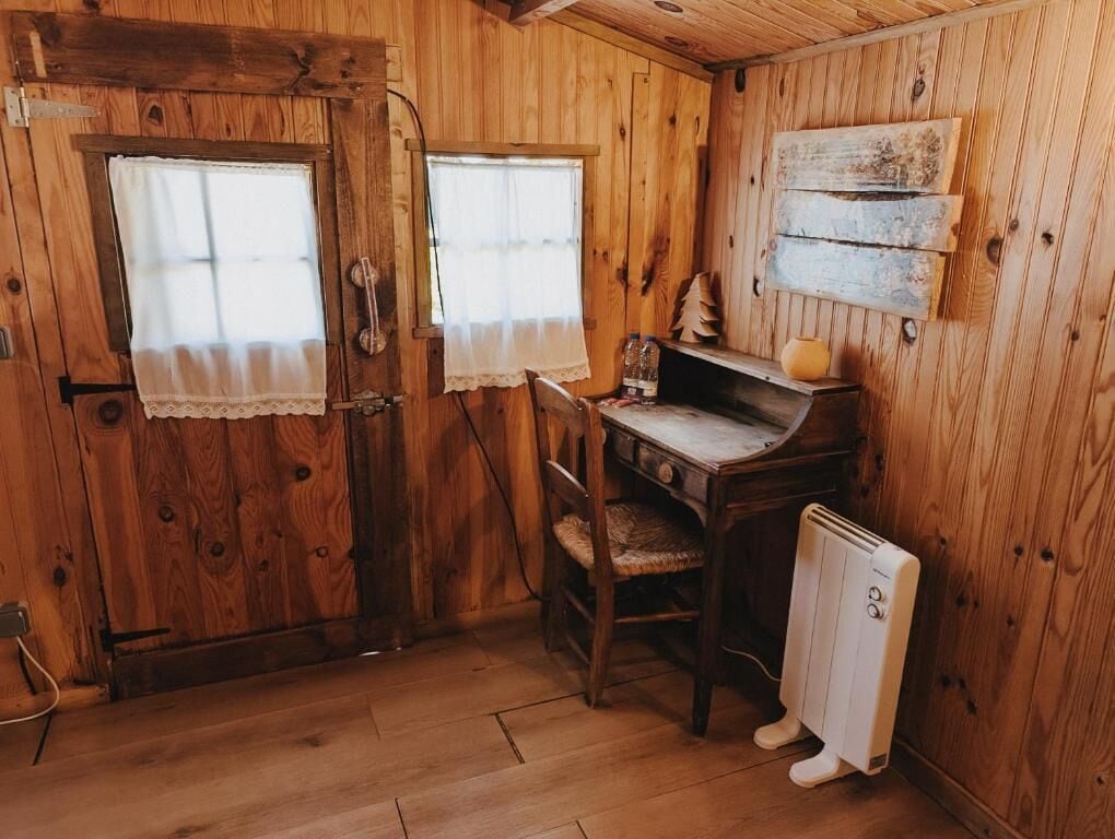 A wooden room with a desk, chair, radiator, and two windows with curtains.