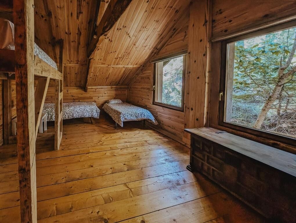 A cabin with a sloped ceiling, two bunk beds, a window with a view of trees, and a fireplace.
