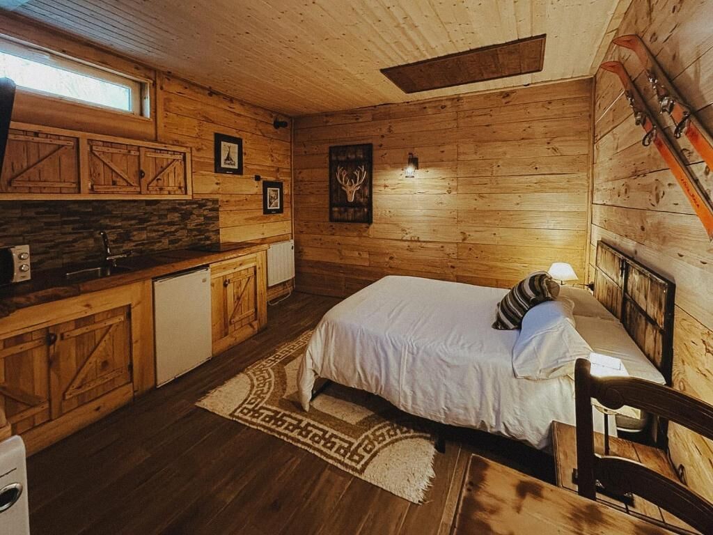 Interior de una cabaña de madera con una cama, una pequeña kitchenette y un soporte para esquíes.