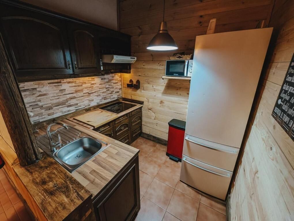Cocina con alacenas de madera oscura, protector contra salpicaduras de piedra, un lavamanos de acero inoxidable, un refrigerador de dos puertas y un microondas sobre una repisa de madera.