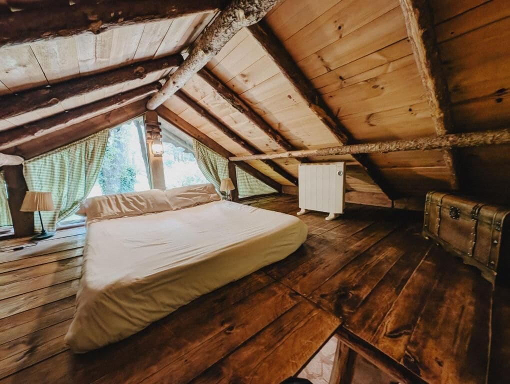 Una habitación con suelo de madera, una cama con ropa blanca, un radiador y una ventana con cortinas.