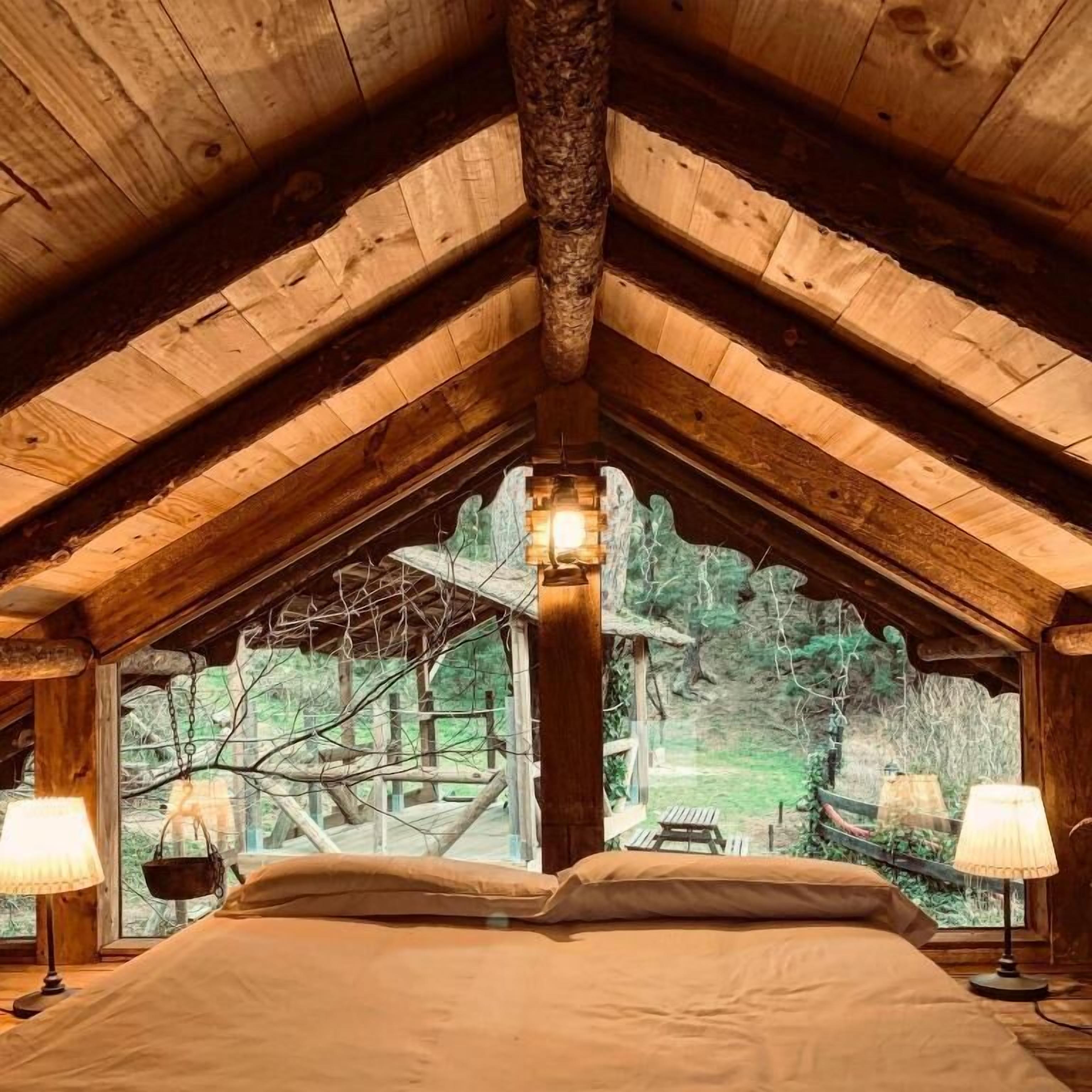 Une chambre avec un plafond en pente, un lit, deux lampes de chevet et une fenêtre donnant sur une zone boisée.