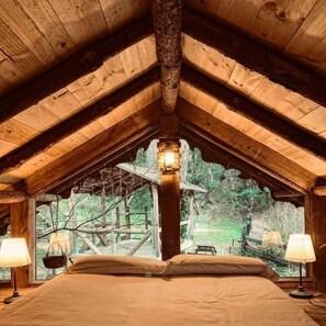A bedroom with a sloped ceiling, a bed, two bedside lamps, and a window with a view of a wooded area.