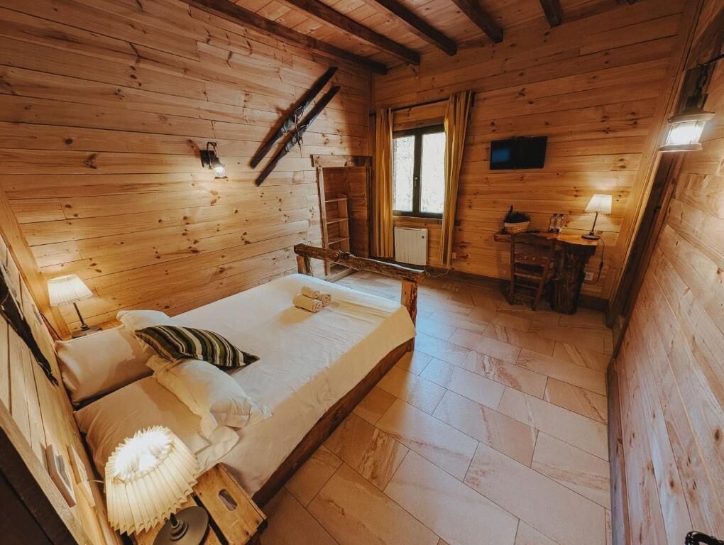 A wooden cabin bedroom with a bed, bedside lamps, a small table, and a window.