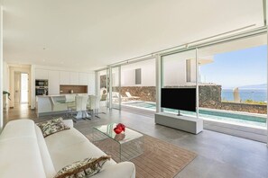 Ein modernes Wohnzimmer mit einer Couch, einem Glastisch und einem Fernseher. Im Hintergrund befindet sich eine Küche und durch große Fenster ist der Meerblick zu sehen.