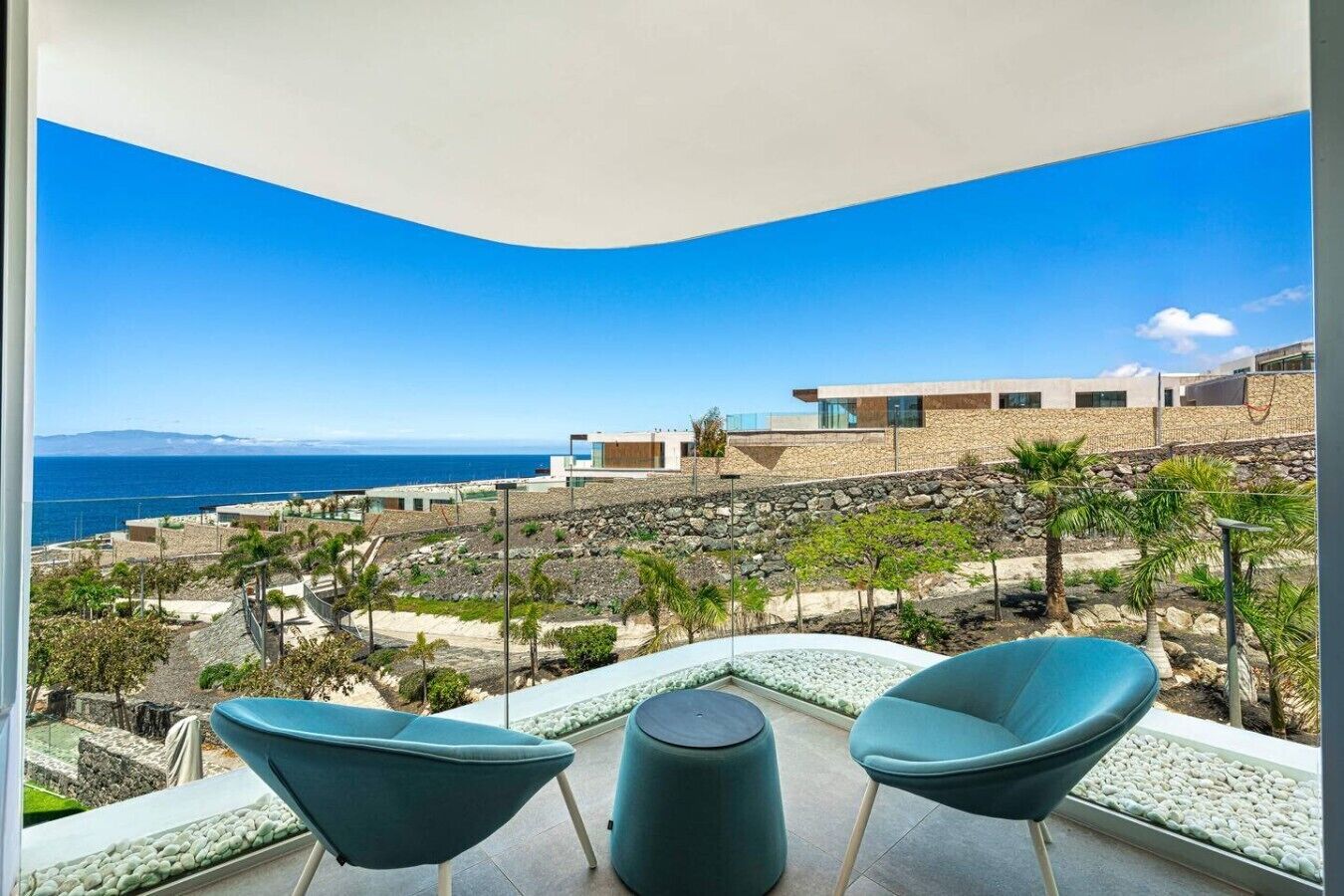 Un balcón con dos sillas y una mesa con vistas a un paisaje costero, edificios modernos y cielo despejado.