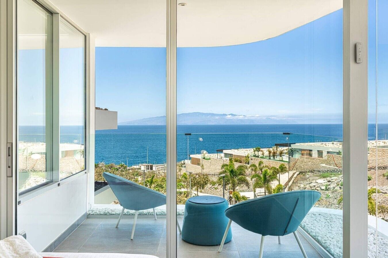 A balcony with a view of the sea and mountains, two blue chairs, and a small round table.