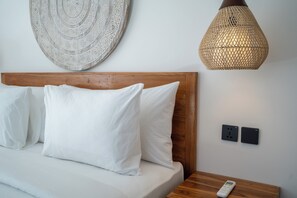 A neatly made bed with white linens, a wooden headboard, a woven pendant light, a wall-mounted power outlet, and a remote control on a wooden nightstand. - Kubu Kaca Cottage (Ungasan)