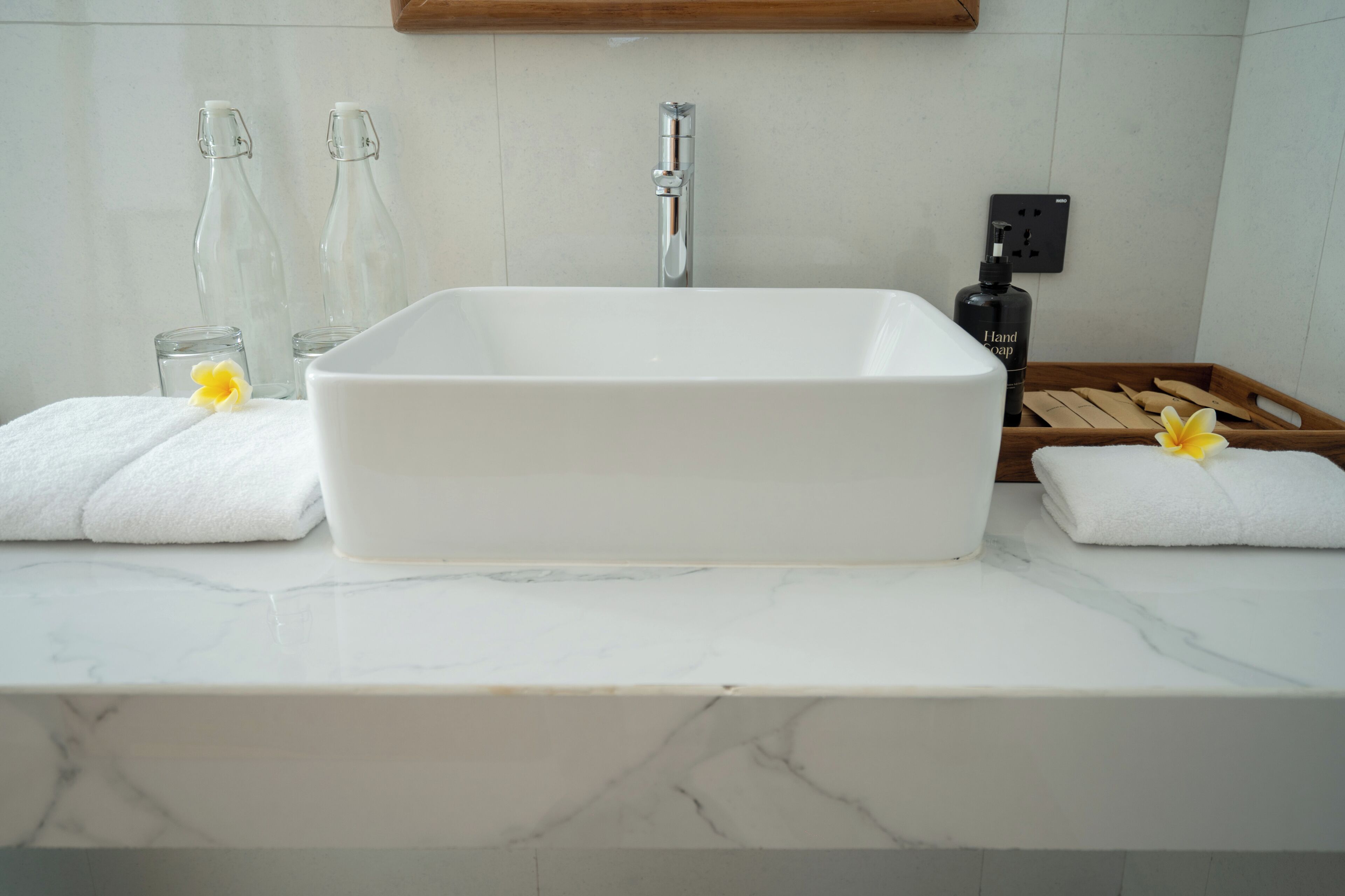 A bathroom with a white rectangular sink, two glass bottles, a faucet, a soap dispenser, and a wooden tray with towels.