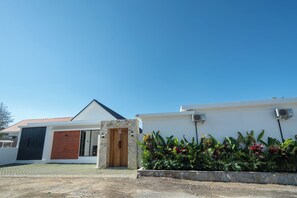 A modern building with a stone facade, a wooden door, and a garden with tropical plants. - Kubu Kaca Cottage (Ungasan)