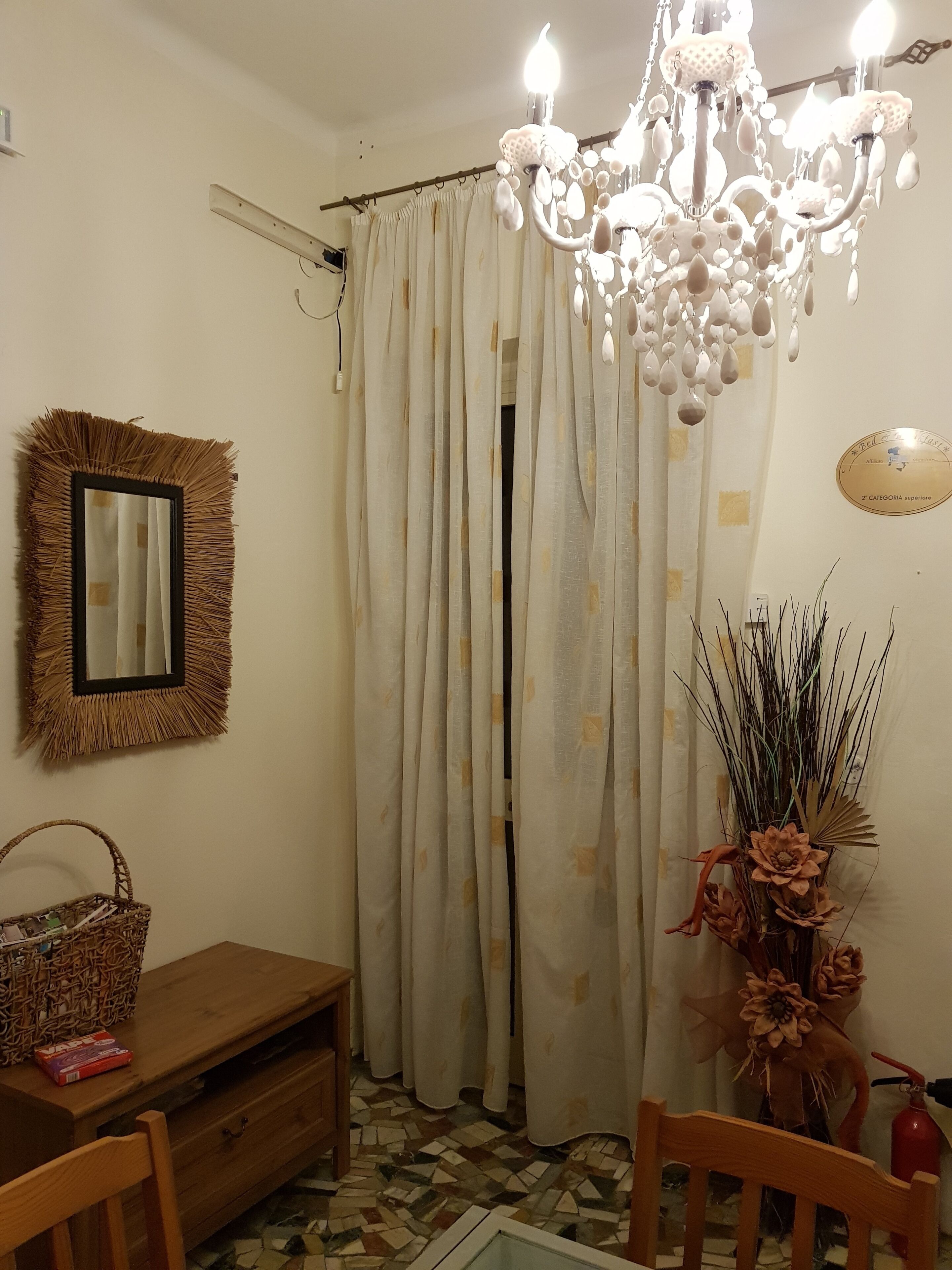 A room with a chandelier, a mirror, a curtain, a vase with dried plants, a wooden table, and a chair.