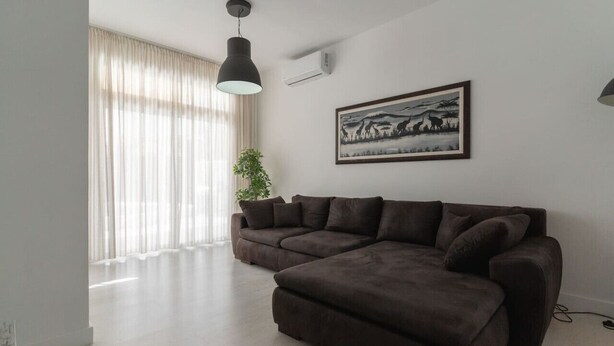 Ein modernes Wohnzimmer mit einem Ecksofa, einem großen Fenster mit durchsichtigen Vorhängen, einer Stehlampe und einem gerahmten Bild an der Wand.
