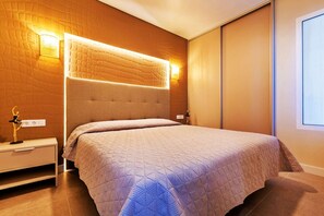 A modern bedroom with a large bed, a headboard with integrated lighting, a nightstand, and a wall-mounted lamp. - 1bedroom ocean view apartment (Costa Adeje)