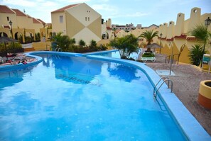 Ein Swimmingpool mit glasklarer, blauer Wasseroberfläche, umgeben von einer gepflasterten Fläche mit Liegestühlen und Sonnenschirmen.