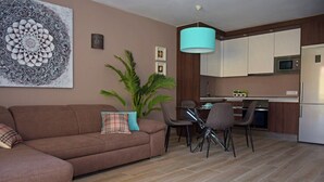 Sala de estar con sofá, mesa de comedor con sillas, zona de cocina con armarios y electrodomésticos, y un gran elemento decorativo en la pared.