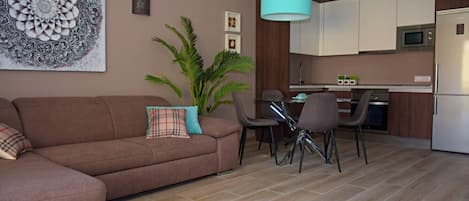 Sala de estar con sofá, mesa de comedor con sillas, zona de cocina con armarios y electrodomésticos, y un gran elemento decorativo en la pared.