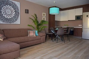 Ein Wohnzimmer mit einer Couch, einem Esstisch mit Stühlen, einer Küchenzeile mit Schränken und Geräten sowie einem großen Wandbild.
