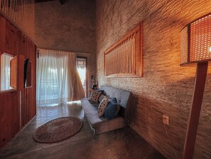 A cozy room with a wooden floor, a grey sofa with patterned cushions, a woven rug, and a wall-mounted lamp. - Quarto triplo beira mar (Cruz)