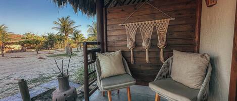 A wooden chair and a cushioned bench outside a building with a thatched roof, a macrame wall hanging, and a view of palm trees and a beach.