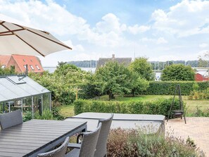 A patio with a table and chairs, a glass greenhouse, and a view of a lake and buildings in the distance. - 4 Star Holiday Home in Nysted (Nysted)