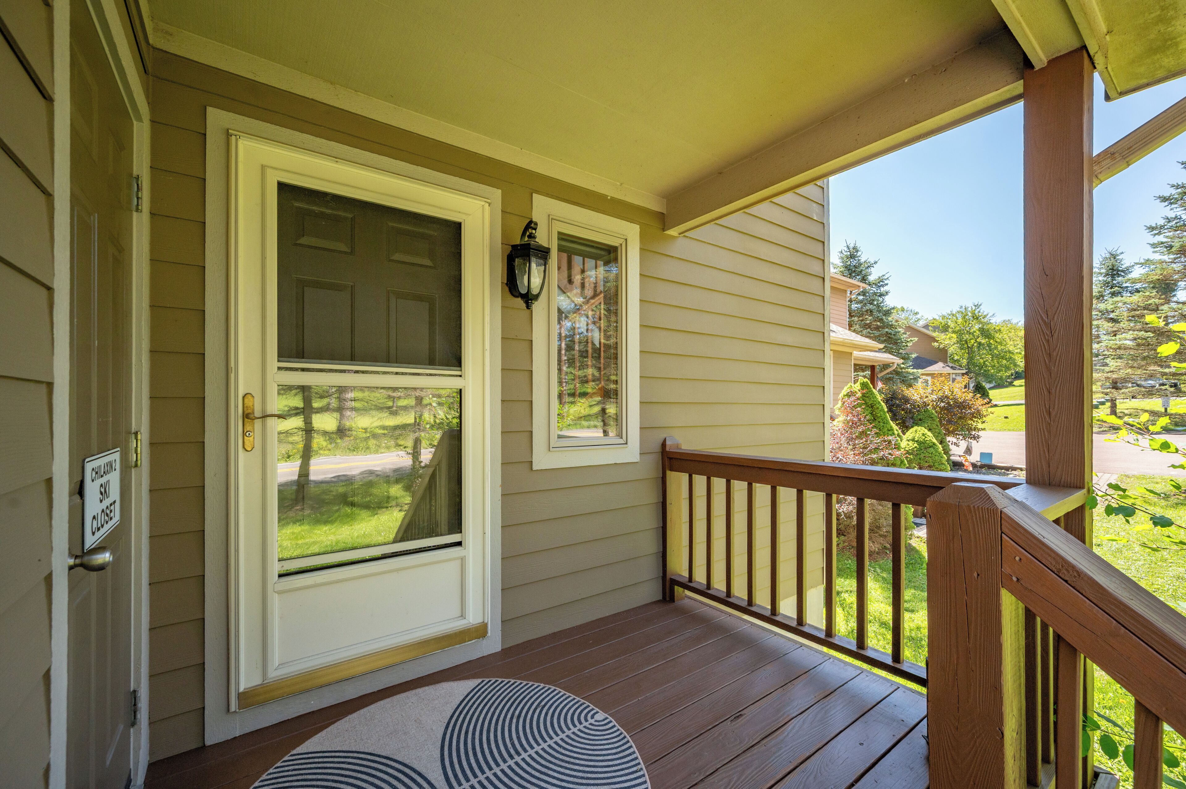 Eine hölzerne Veranda mit einer weißen Tür und einem kleinen Fenster, einem Teppich und Blick auf eine grüne Landschaft.