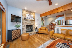 A living room with a stone fireplace, wooden beams, a TV, and a sofa. - New! Lake Area Townhome w/ Hot Tub (McHenry)