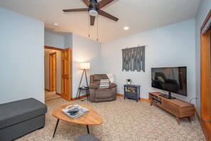 A living room with a ceiling fan, a television, a sofa, a coffee table, and a fireplace. - New! Lake Area Townhome w/ Hot Tub (McHenry)