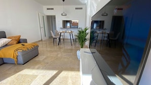 A modern living room with a sofa, dining table, and chairs, a kitchen area, and a large window. - Ocean-View Apartment with Pool in Los Gigantes (Santiago del Teide)