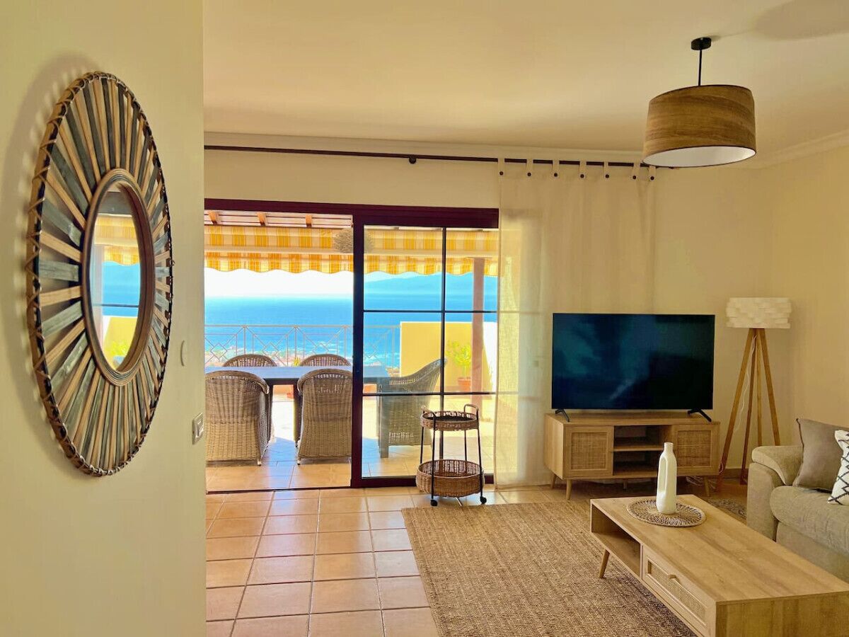 A living room with a television, a sofa, a coffee table, and a view of the sea through large glass doors.