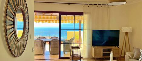 A living room with a television, a sofa, a coffee table, and a view of the sea through large glass doors.