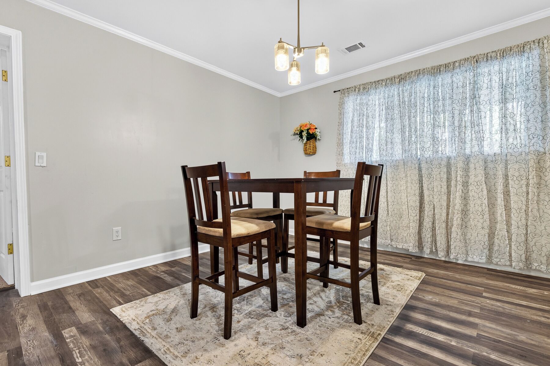 A dining area with a round table and four chairs, a chandelier, a vase with flowers, and patterned curtains.