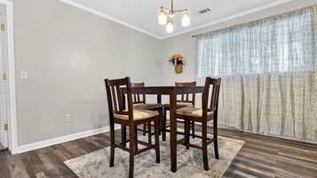 A dining area with a round table and four chairs, a chandelier, a vase with flowers, and patterned curtains.
