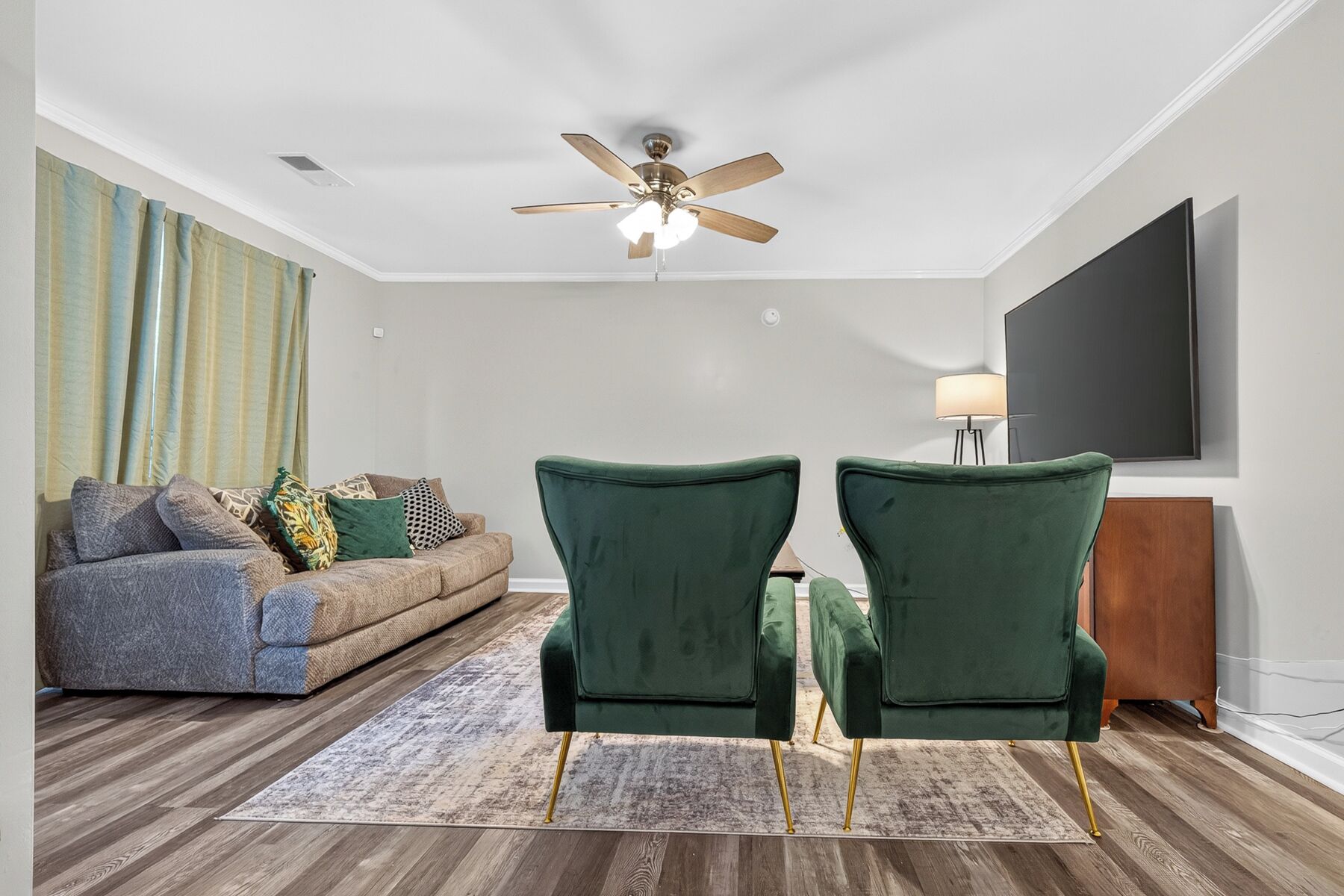 A living room with a ceiling fan, a sofa, two green armchairs, a rug, and a flat-screen TV mounted on the wall.