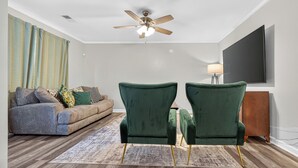 A living room with a ceiling fan, a sofa, two green armchairs, a rug, and a flat-screen TV mounted on the wall.