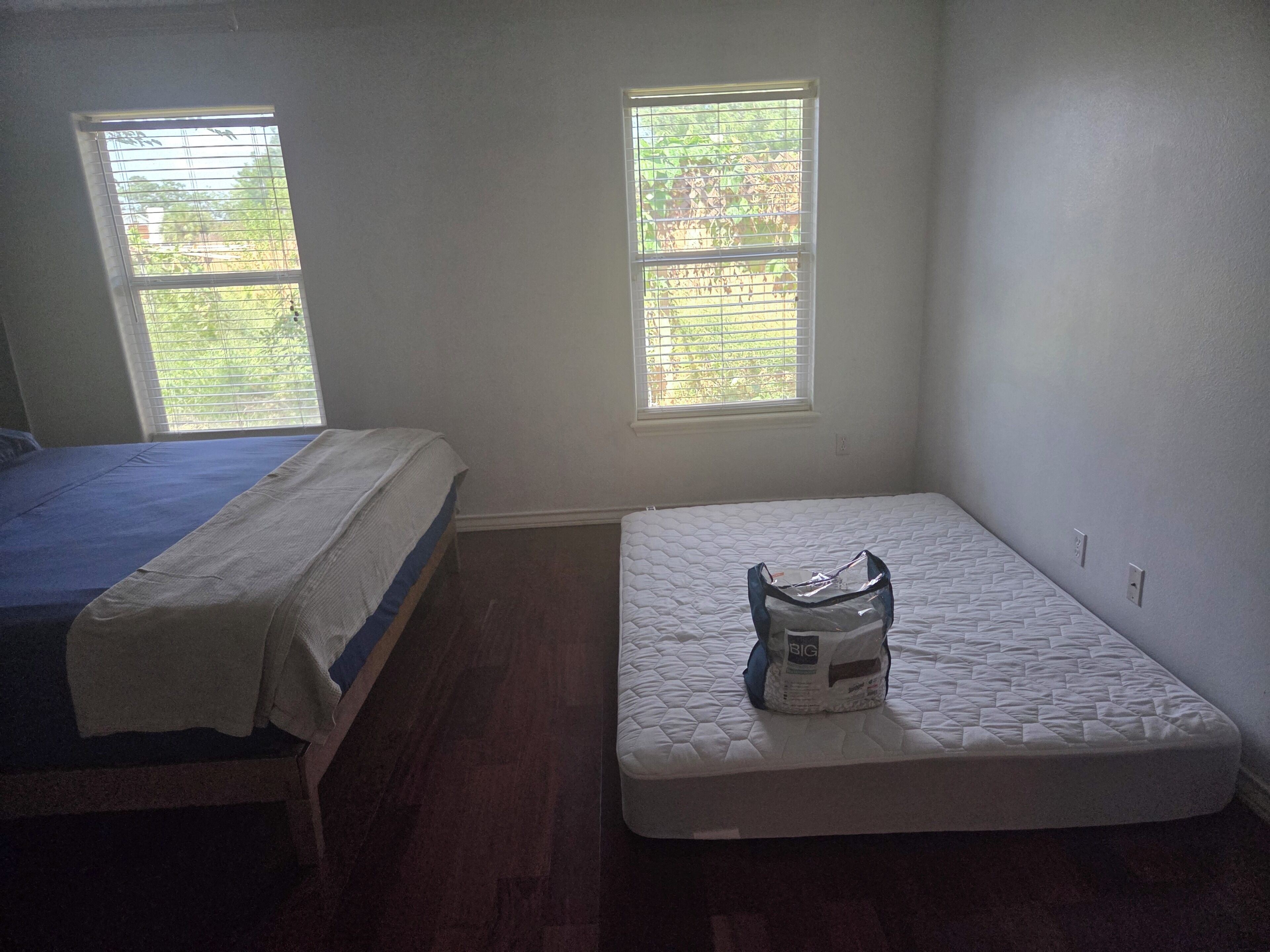 A room with two beds, one with a blue bedspread and the other with a white bedspread. There are two windows with blinds, and the room has a wooden floor.