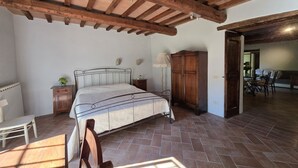 A bedroom with a bed, wooden headboard, a nightstand, a lamp, a wardrobe, and a dining area in the background. - Villa Tufi by MMega (Montone)